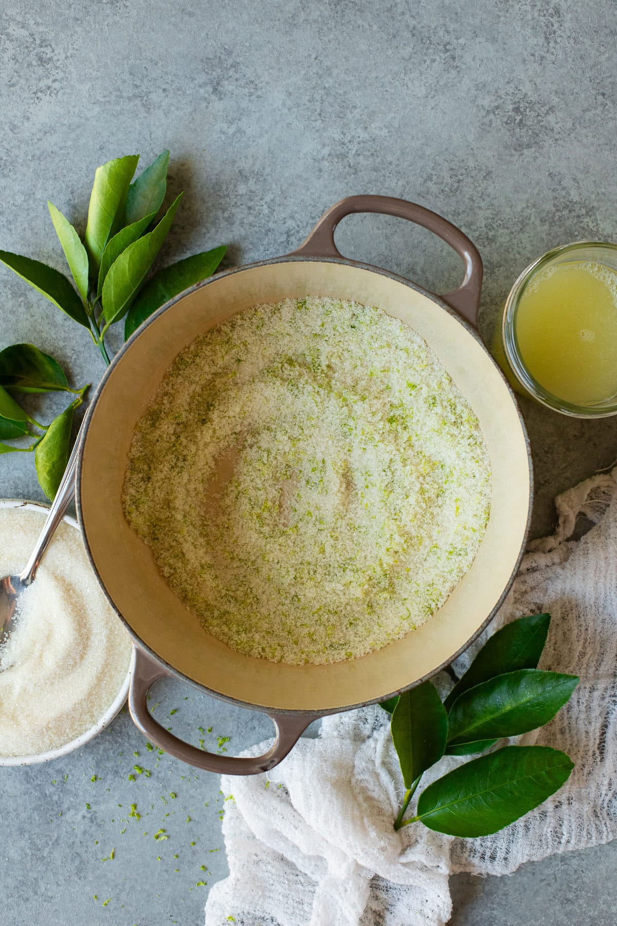 Lime zest and granulated sugar mixed together in a pot, alongside a shallow bowl of sugar and a jar of lime juice.